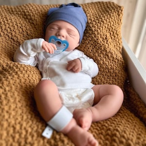 Puede incluir: Una mu&ntilde;eca beb&eacute; realista con gorro azul, body blanco y pa&ntilde;al, con un chupete azul en la boca. La mu&ntilde;eca est&aacute; acostada sobre una manta de punto marr&oacute;n. Una banda blanca envuelve la pierna del beb&eacute;.
