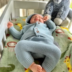 Puede incluir: Un beb&eacute; con un c&aacute;rdigan con capucha y pantalones de punto azul claro yace en una cuna blanca. El beb&eacute; est&aacute; sobre una manta verde con dise&ntilde;os de animales. Un elefante de peluche gris est&aacute; en el fondo.
