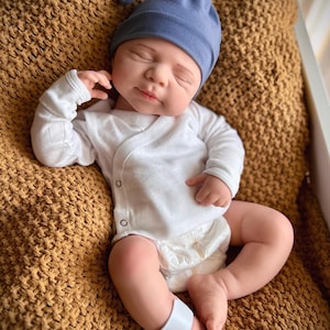 Puede incluir: Una mu&ntilde;eca beb&eacute; realista con un gorro azul anudado, un body blanco cruzado y un pa&ntilde;al. La mu&ntilde;eca est&aacute; acostada sobre una manta marr&oacute;n texturizada. Una banda blanca est&aacute; alrededor de la pierna de la mu&ntilde;eca.