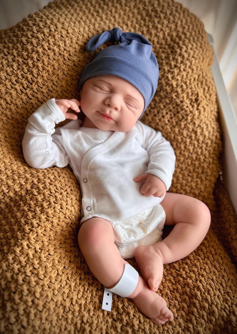 Puede incluir: Una mu&ntilde;eca beb&eacute; realista con un gorro azul anudado, un body blanco cruzado y un pa&ntilde;al. La mu&ntilde;eca est&aacute; acostada sobre una manta marr&oacute;n texturizada. Una banda blanca est&aacute; alrededor de la pierna de la mu&ntilde;eca.