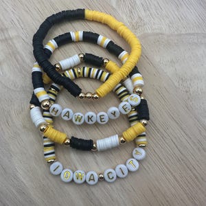 May include: Set of four bracelets with black and yellow beads. The bracelets spell out "HAWKEYES" and "IOWA CITY" in gold letters.