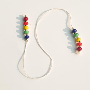 May include: A handmade rainbow bead garland with red, orange, yellow, green, and blue beads. The beads are strung on a natural-colored cord, with gold-colored accents at each end. The garland is displayed on a white background.