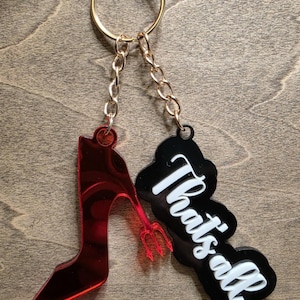 May include: A keychain featuring a red high-heeled shoe and a black tag with the text "That's all" in white. The shoe has a devil's trident. The keychain is attached to a gold ring with a gold chain.