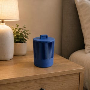 Könnte beinhalten: Ein zylindrischer blauer Behälter mit gerippter Textur in der oberen Hälfte und einem flachen Deckel, der oben einen kleinen rechteckigen Griff aufweist. Das Objekt hat einen festen Boden und eine gleichmäßige matte Oberfläche.