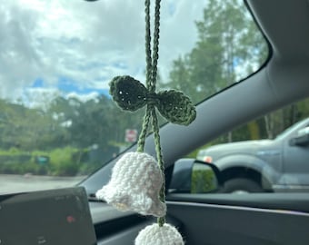 May include: A handmade, crochet car charm featuring two white bell-shaped flowers suspended from a green stem and bow. The charm is hanging from a car's rearview mirror, with a blurred background of trees and a cloudy sky.