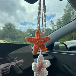 Könnte beinhalten: Ein handgefertigter Autoanhänger mit einem gehäkelten orangefarbenen Seestern und einer weißen Blume mit rosa Akzenten. Der Anhänger hängt an einer Kette aus Muscheln.