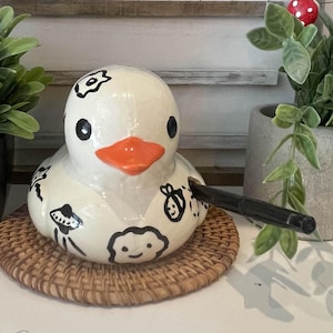 May include: A ceramic duck figurine with black and white whimsical designs, an orange beak, and black eyes. The duck is on a woven mat, with the text "TOADSTOOL CERAMICS" in the background.