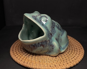 May include: A ceramic frog figurine with a wide open mouth, painted in shades of teal and brown. The frog is sitting on a woven wicker coaster.