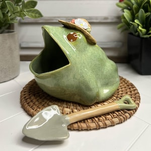 Może przedstawiać: Zielona ceramiczna donica w kształcie żaby z otwartą paszczą, w małym kapeluszu. Pasująca łopatka spoczywa na plecionej macie. W tle widnieje napis "TOADSTOOL CERAMICS".