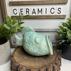 May include: A teal blue ceramic fish with a textured surface and a large open mouth. The fish is sitting on a wooden log slice. The sign behind the fish reads "Toadstool Ceramics".