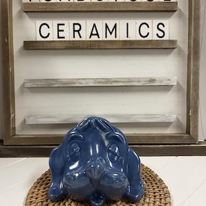 May include: A blue ceramic dog head figurine with a whimsical expression. The figurine is sitting on a woven wicker coaster. The background features a sign that reads "Toadstool Ceramics".