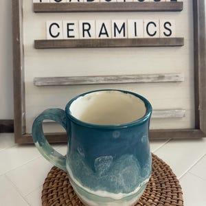 May include: A teal blue ceramic mug with a white and blue wave design. The mug is sitting on a woven coaster. The mug has a handle and is a handmade pottery piece. The mug is labelled "Toadstool Ceramics".