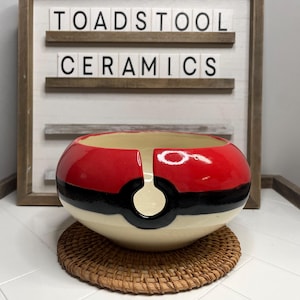 May include: A ceramic yarn bowl with a red, black, and white Pokeball design. The bowl has a hole in the center for yarn to be dispensed.
