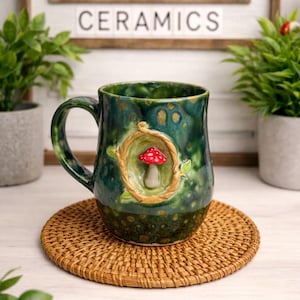 May include: A green ceramic mug with a mushroom design. The mug has a handle and is decorated with a red and white mushroom inside a circular frame. The mug sits on a woven coaster. The sign behind the mug says "TOADSTOOL CERAMICS."