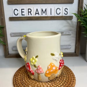 Könnte beinhalten: Cremefarbene Keramik-Tasse mit Henkel, verziert mit bunten Pilz- und Blumenapplikationen. Die Tasse steht auf einem runden Untersetzer aus Weide. Der Text "TOADSTOOL CERAMICS" ist im Hintergrund zu sehen.