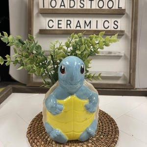 May include: A blue and yellow ceramic turtle planter with green foliage, set on a round woven mat. The sign behind the planter reads "TOADSTOOL CERAMICS". The planter is a decorative item.