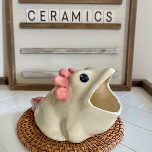 May include: A white ceramic frog with pink accents and a large open mouth. The frog is sitting on a woven brown wicker coaster. The frog is a vintage style kitchen utensil holder.