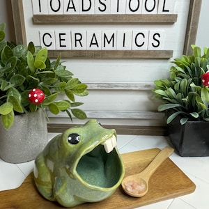 May include: Green ceramic frog salt dish with a toothy grin, sitting on a wooden chopping board with a small wooden spoon and pink Himalayan salt. The frog is glazed in a light green colour and has black eyes. The words "Toadstool Ceramics" are displayed on a white sign behind the frog.