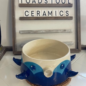May include: A handmade ceramic bowl with a whimsical design. The bowl features a cream-colored interior and is decorated with shades of blue, resembling a creature. The text "TOADSTOOL CERAMICS" is displayed in the background.