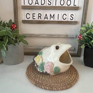 Könnte beinhalten: Ein weißer Keramikbehälter in Fischform mit offenem Maul, verziert mit gelben, rosa und grünen Blumen, steht auf einem gewebten Untersetzer. Das Schild dahinter lautet "TOADSTOOL CERAMICS."