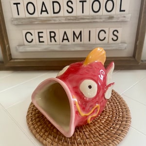 Könnte beinhalten: Ein Keramik-Pflanztopf in Fischform mit rosa Glasur und gelben Akzenten. Der Topf hat eine große Öffnung zum Pflanzen. Der Topf steht auf einem geflochtenen Untersetzer.