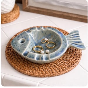 May include: A blue ceramic fish-shaped trinket dish with a textured finish. The dish holds several gold rings and two small pearl earrings. It rests on a round, woven rattan coaster, suggesting a coastal or nautical theme.