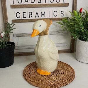 May include: A ceramic duck ornament with a cream body, orange beak and feet, and a brown neck piece. The duck sits on a round, woven, brown placemat. The text "TOADSTOOL CERAMICS" is visible in the background.