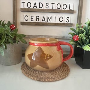 Può includere: Una tazza in ceramica con base beige, manico rosso e design a stella marrone. La tazza è appoggiata su un sottobicchiere intrecciato. L'insegna dietro la tazza recita "TOADSTOOL CERAMICS".