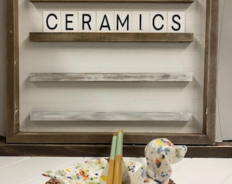 May include: A white ceramic dog-shaped dish with colourful speckles, holding two wooden chopsticks with green tips. The dish is sitting on a woven wicker coaster. A wooden sign with the words "Toadstool Ceramics" is in the background.