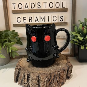 Könnte beinhalten: Schwarze Keramik-Tasse mit roten augenartigen Akzenten und spitzenartigen Details. Die Tasse hat einen Henkel und wird auf einer Holzscheibe präsentiert. Der Text "TOADSTOOL CERAMICS" befindet sich im Hintergrund.