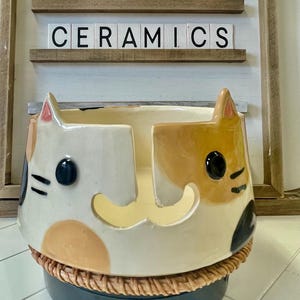 May include: Ceramic yarn bowl featuring a cat design. The bowl is white with brown, black and orange accents. The bowl has a woven base.