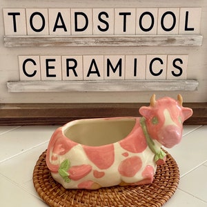 Puede incluir: Una maceta de cerámica con forma de vaca, con un patrón de fresas rosas y blancas, sobre una estera tejida. La maceta tiene pequeños cuernos y un detalle de hoja verde. El texto "TOADSTOOL CERAMICS" se muestra arriba.