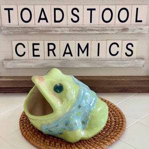 May include: A green ceramic frog figurine with a blue and white floral pattern. The frog has a large open mouth, making it suitable for use as a dish or trinket holder. The frog is sitting on a woven wicker coaster. The sign above reads "Toadstool Ceramics".
