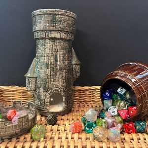 以下が含まれることがあります： 緑色の屋根が付いたセラミック製の塔と、蓋が付いた茶色の樽があり、どちらもカラフルなサイコロでいっぱいです。サイコロは籐のバスケットの上に散らばっています。