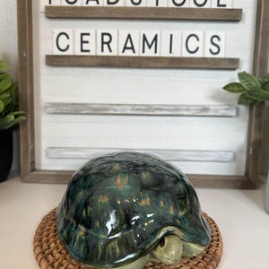Puede incluir: Una figura de tortuga de cerámica con un esmalte brillante moteado verde y dorado, sobre un posavasos tejido. El letrero detrás dice "TOADSTOOL CERAMICS". La cabeza de la tortuga es visible, agregando detalles a la pieza artesanal.