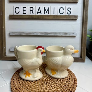 May include: Two ceramic egg cups, one shaped like a chicken and the other like a duck. Both are white with yellow feet and beaks, and the chicken has a red comb. The egg cups are sitting on a woven wicker coaster.
