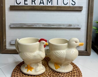 Puede incluir: Dos soportes para huevos de cerámica, uno con forma de pollo y el otro con forma de pato. Ambos son blancos con patas y picos amarillos, y el pollo tiene una cresta roja. Los soportes para huevos están sobre un posavasos de mimbre tejido.