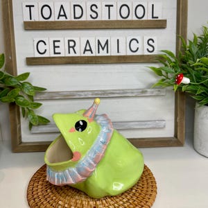 Könnte beinhalten: Ein skurriler, limettengrüner Keramik-Blumentopf in Form einer Kreatur mit Rüschenkragen. Der Topf steht auf einer gewebten, kreisförmigen Matte. Der Text "TOADSTOOL CERAMICS" ist im Hintergrund zu sehen.