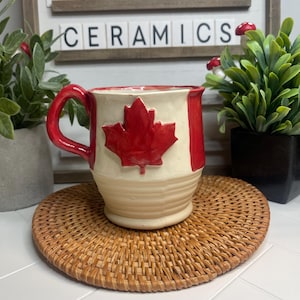 May include: A white ceramic mug with a red Canadian flag design. The mug has a red handle and a white background with a red maple leaf in the center. The mug is sitting on a woven brown coaster.