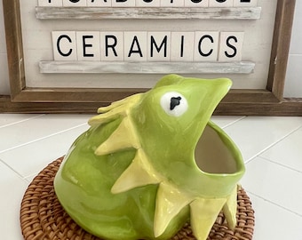 May include: A green ceramic frog-shaped dish with a wide open mouth. The dish is sitting on a woven brown wicker coaster. The dish is labeled "Toadstool Ceramics".