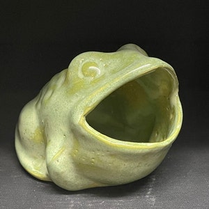 Può includere: Un contenitore a forma di rana in ceramica nei toni del verde e giallo. La rana ha la bocca aperta, formando una forma a ciotola. Il contenitore è probabilmente per uso decorativo o funzionale.