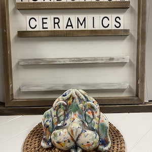 May include: A ceramic dog head sculpture with a colorful, speckled glaze. The sculpture is resting on a woven wicker coaster. The background features a wooden sign with the words "Toadstool Ceramics" in white letters.