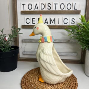 May include: A cream-coloured ceramic goose planter with a yellow beak and feet, wearing a colourful collar and party hat. The planter sits on a woven mat. The text "TOADSTOOL CERAMICS" is in the background.