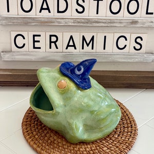 Könnte beinhalten: Eine grüne Keramik-Frosch mit einem blauen Zaubererhut, der auf einem geflochtenen braunen Weidenuntersetzer sitzt. Der Frosch hat einen großen, offenen Mund und der Hut hat einen weißen Kreis mit einem Stern in der Mitte. Der Frosch ist ein Pflanztopf oder eine Schale.