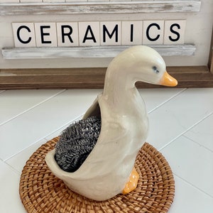 May include: A ceramic goose-shaped holder with a gray scrubby inside, resting on a round woven mat. The goose is off-white with an orange beak and feet. The text "TOADSTOOL CERAMICS" is displayed above.