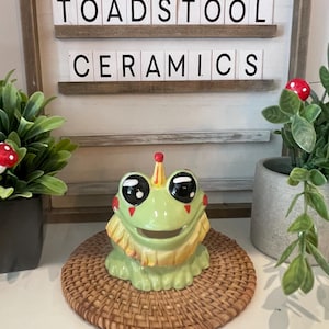 May include: A green ceramic frog figurine with large black eyes, red accents, and a yellow crown, sitting on a woven mat. The sign behind it reads "TOADSTOOL CERAMICS". Decorative plants with red mushroom accents are on either side.
