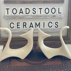 Op de afbeelding: Twee witte keramische sculpturen van gestileerde katten, die elkaar aankijken. De sculpturen staan op een bruin, gepatroneerd oppervlak. Op de achtergrond is een bord te zien met de tekst "Toadstool Ceramics".