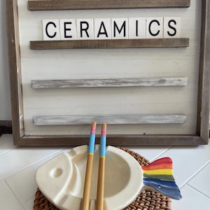 May include: White ceramic fish-shaped dish with a rainbow tail, sitting on a woven mat. Two wooden chopsticks with blue and pink accents are placed inside the dish. The dish is labelled "Toadstool Ceramics" on a wooden sign behind it.