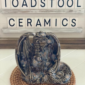 Peut inclure: Une sculpture de dragon en céramique aux ailes déployées, dans des tons de bleu, marron et or, posée sur un tapis tressé. Le texte "TOADSTOOL CERAMICS" est affiché au-dessus de la sculpture sur un fond blanc.