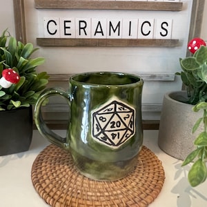 Könnte beinhalten: Grüne Keramiktasse mit einem schwarz-weißen D20-Design. Die Tasse hat einen Henkel und steht auf einem gewebten Untersetzer. Der Text "TOADSTOOL CERAMICS" ist im Hintergrund zu sehen.